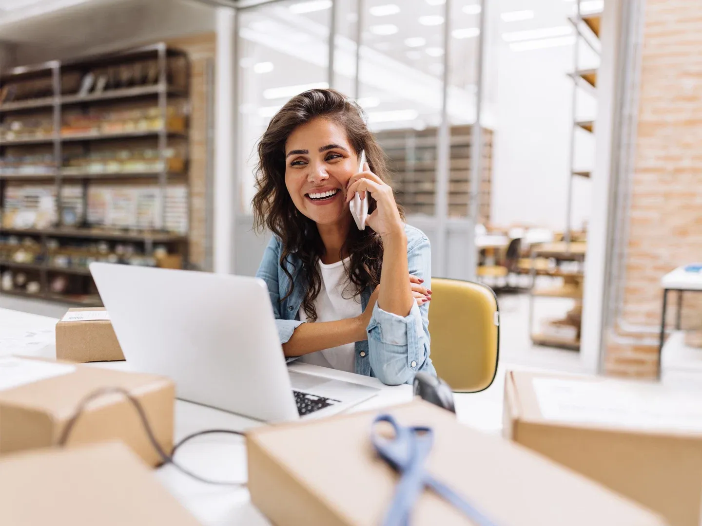 A smiling woman speaking on the phone at a desk with a laptop inside her small business office/warehouse space.